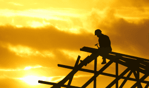 Roofing contractor worker inspecting wooden roof structure at sunset with dramatic sky in background.